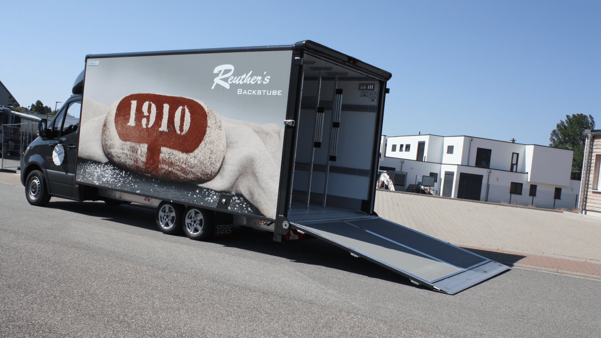 Ein schwarzer Lieferwagen mit offener Heckrampe, auf dessen Seite das Bild eines Brotlaibs mit der Aufschrift 1910 und der Text Reuthers Backstube zu sehen ist, parkt auf einer sonnigen Straße in der Nähe moderner Gebäude.