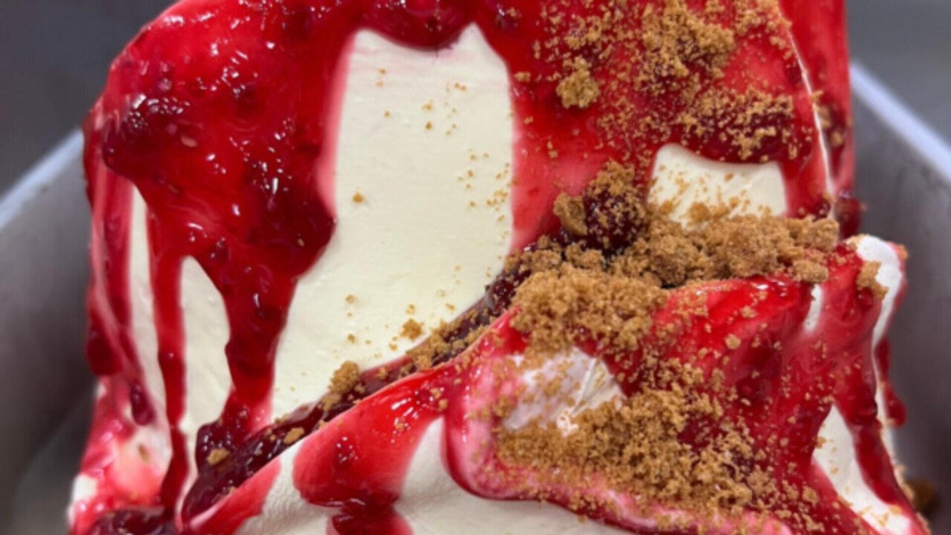 A close-up of creamy vanilla ice cream topped with a vibrant red raspberry sauce and sprinkled with crumbled brown biscuits.