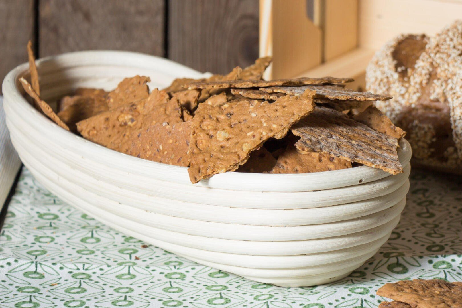 A white, oval-shaped basket filled with crispy brown flatbread crackers sits on a patterned tablecloth, with a loaf of seeded bread in the background.