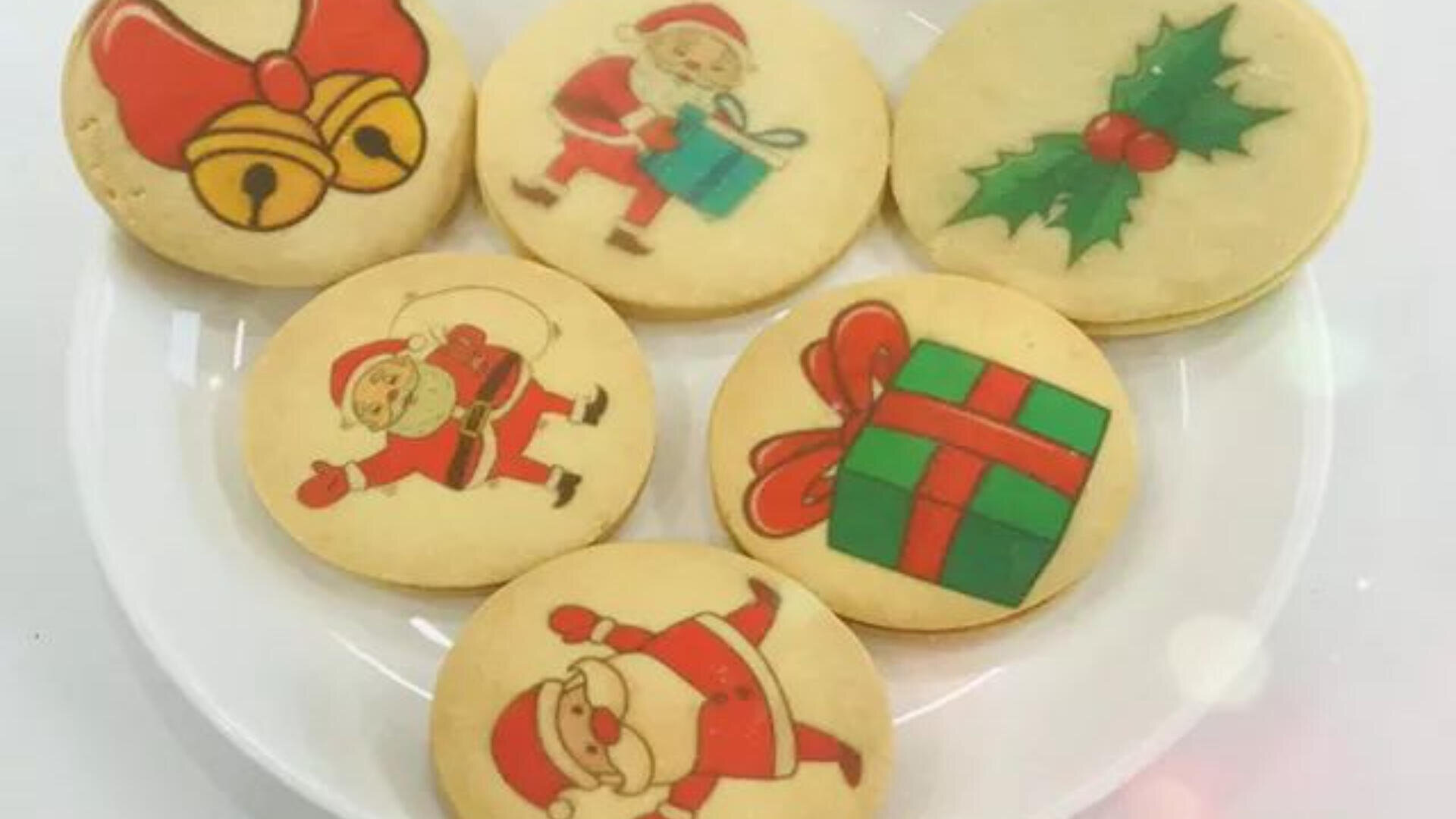 A plate with six decorated cookies featuring Christmas designs: three Santas, a present, holly leaves with berries, and golden bells with a red bow.
