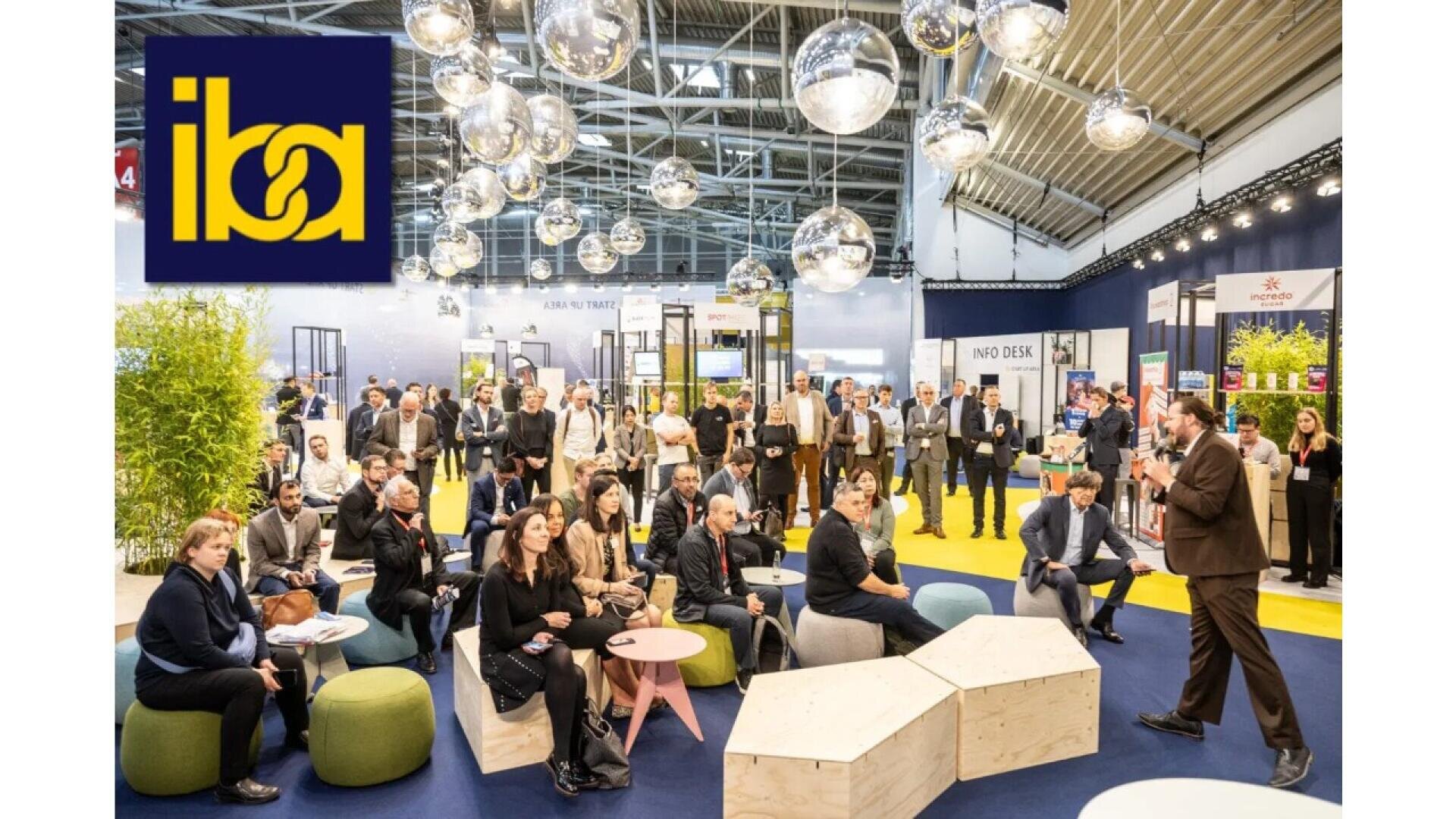 A speaker addresses a seated audience at a modern indoor event with globe lights, yellow carpet, and display booths. The iba logo is visible in the top left corner.
