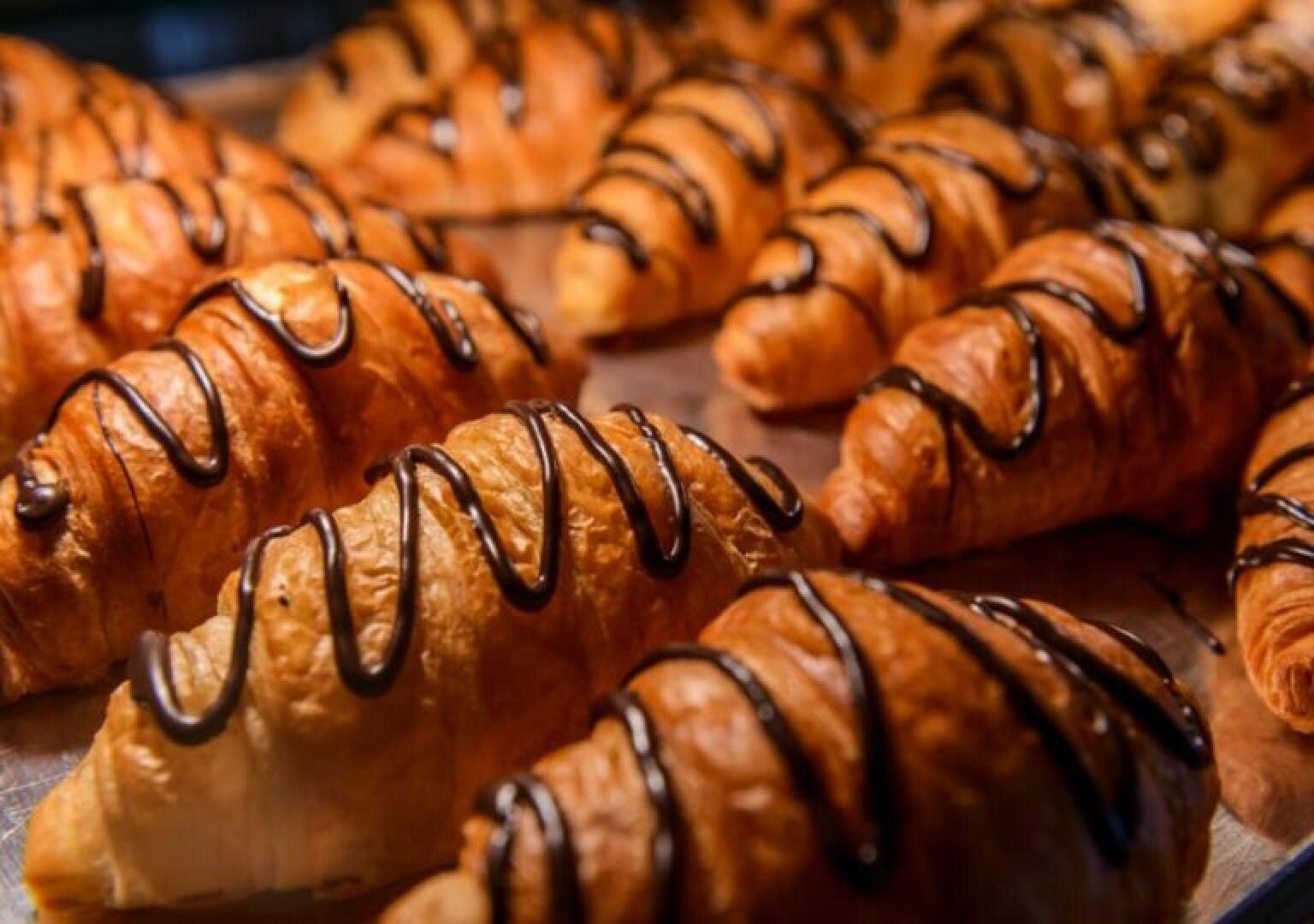 Golden, flaky croissants topped with drizzled chocolate are arranged closely together on a baking tray, creating a tempting and delicious display of pastries.