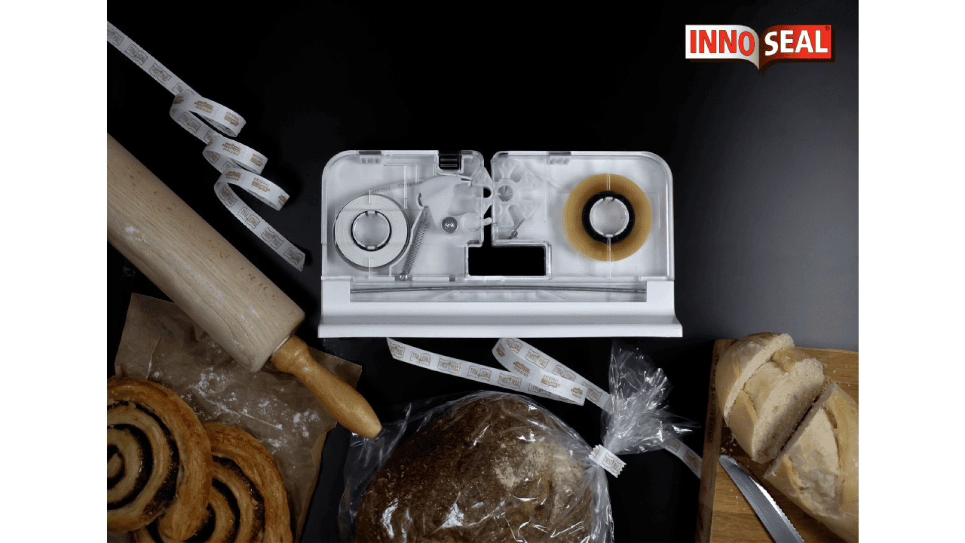 Eine Tischmaschine zum Verschließen von Tüten mit Klebeband und Papierstreifen steht auf einer schwarzen Fläche, umgeben von Broten, einem Nudelholz und Gebäck. In der rechten oberen Ecke ist das Logo "INNOSEAL" zu sehen.
