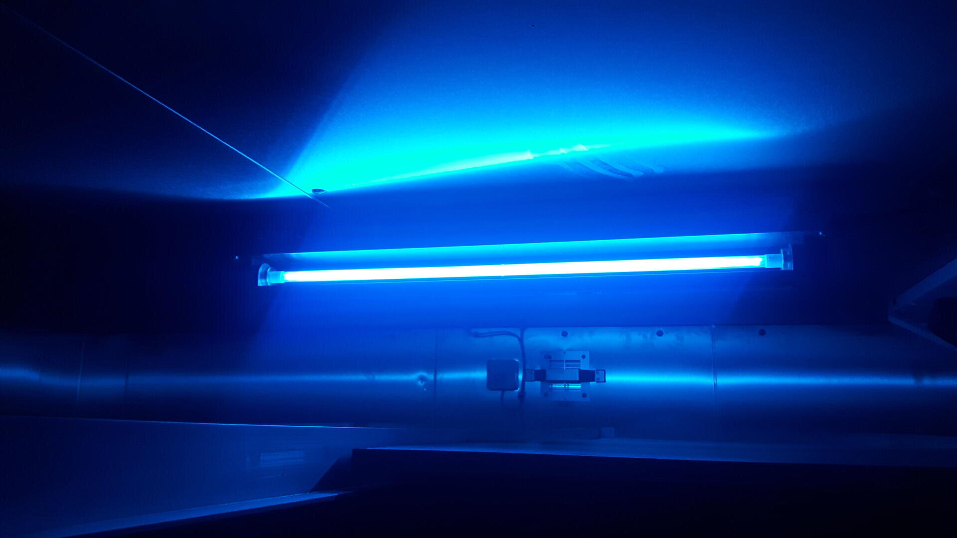 Eine hellblaue UV-Leuchtstoffröhre ist an einer Wand montiert und erhellt die Umgebung mit blauem Licht. Der Raum besticht durch metallische Oberflächen und ein modernes, industrielles Erscheinungsbild.
