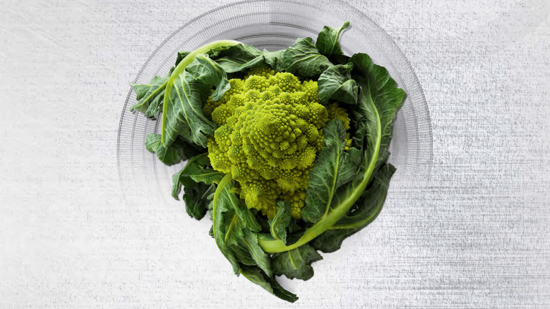 Ein Kopf von Romanesco-Brokkoli mit seinen grünen Blättern auf einer klaren Glasplatte vor einem hellen, strukturierten Hintergrund, von oben betrachtet.