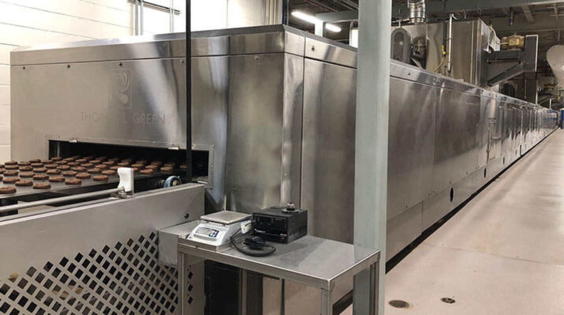 Ein großer industrieller Backofen mit Keksen auf einem Förderband, die aus dem Ofen herausfahren. Ein Metalltisch mit einer digitalen Waage und einem schwarzen Gerät steht in einer Fabrikumgebung neben der Maschine.