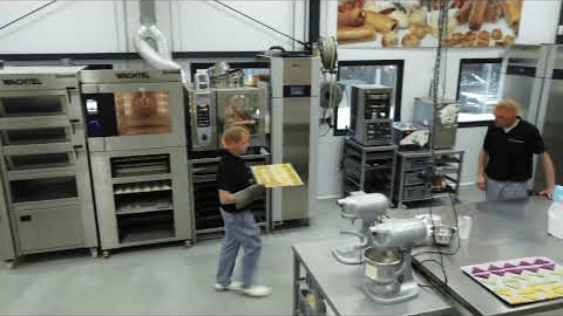 Two people work in a modern bakery kitchen with industrial ovens, mixers, and trays. One person carries a baking tray, while the other stands near stainless steel tables and baking equipment. Baked goods are pictured on the wall.
