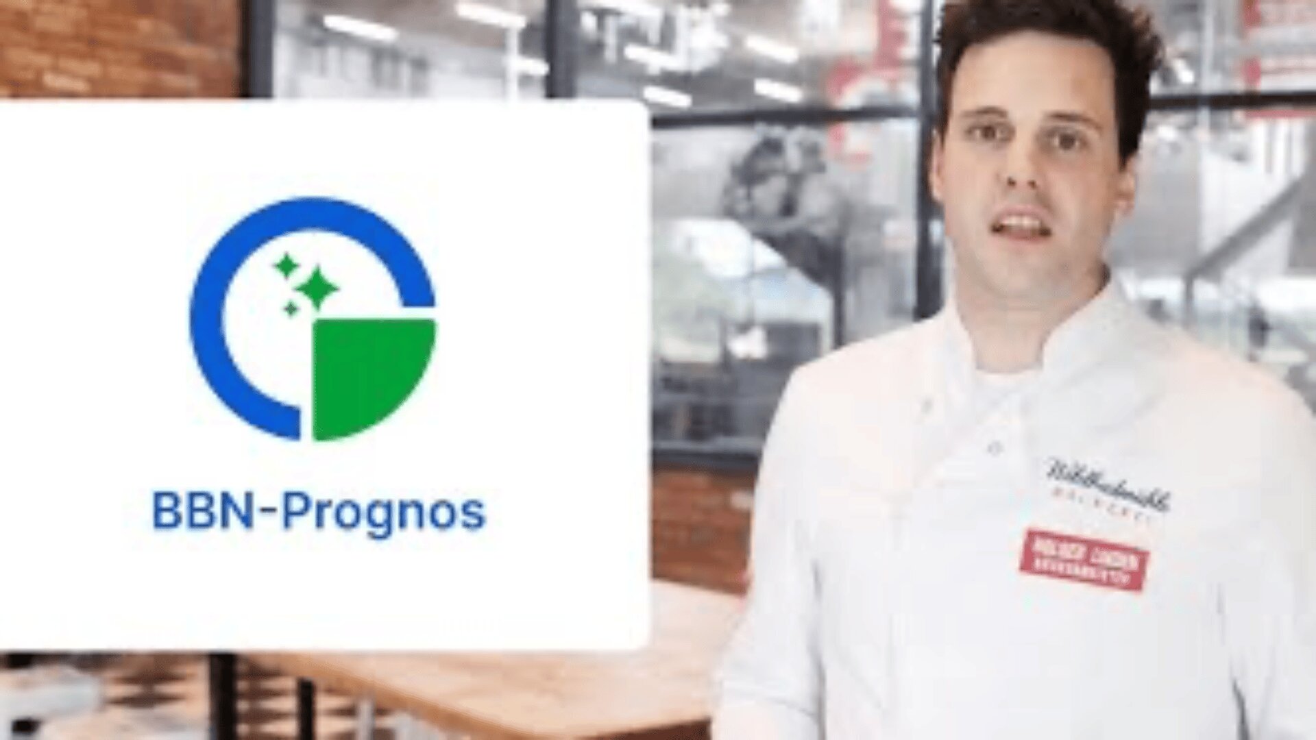 Ein Mann in einer weißen Kochjacke steht in einem Innenraum neben einem Bildschirm, auf dem das Logo von BBN-Prognos in Form eines blauen und grünen Kreises zu sehen ist. Im Hintergrund ist ein modernes Büro mit Glaswänden zu sehen.