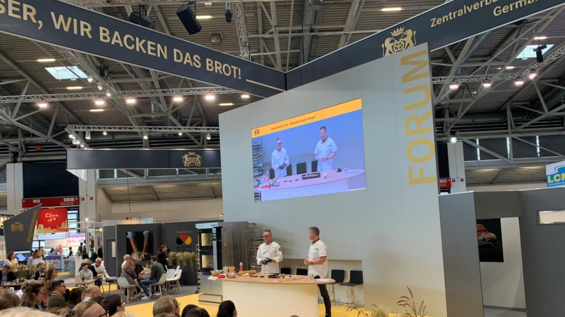 Two men in white shirts give a baking demonstration on stage at a trade show, with an audience watching. A large screen behind them displays their actions. The words “FORUM” and “WIR BACKEN DAS BROT!” are visible above.