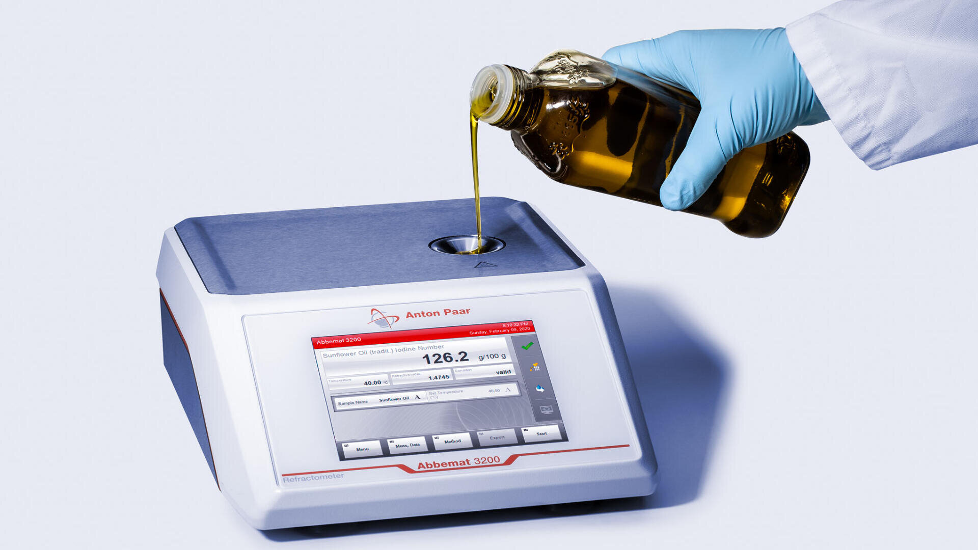A gloved hand pours yellow oil from a bottle into a laboratory device with a digital screen displaying measurement data.