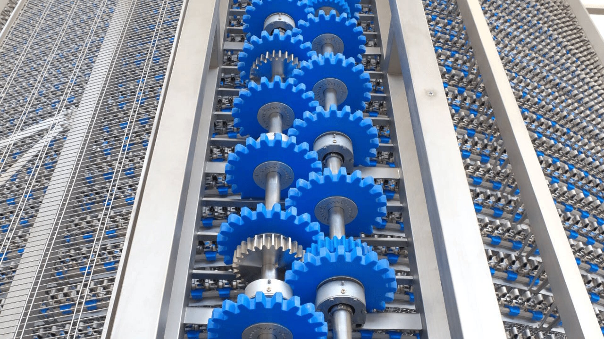 Close-up of a vertical industrial machine with multiple blue gears and metal shafts, surrounded by metal framework and grid panels. The gears are aligned in parallel rows, suggesting a mechanical or manufacturing process.