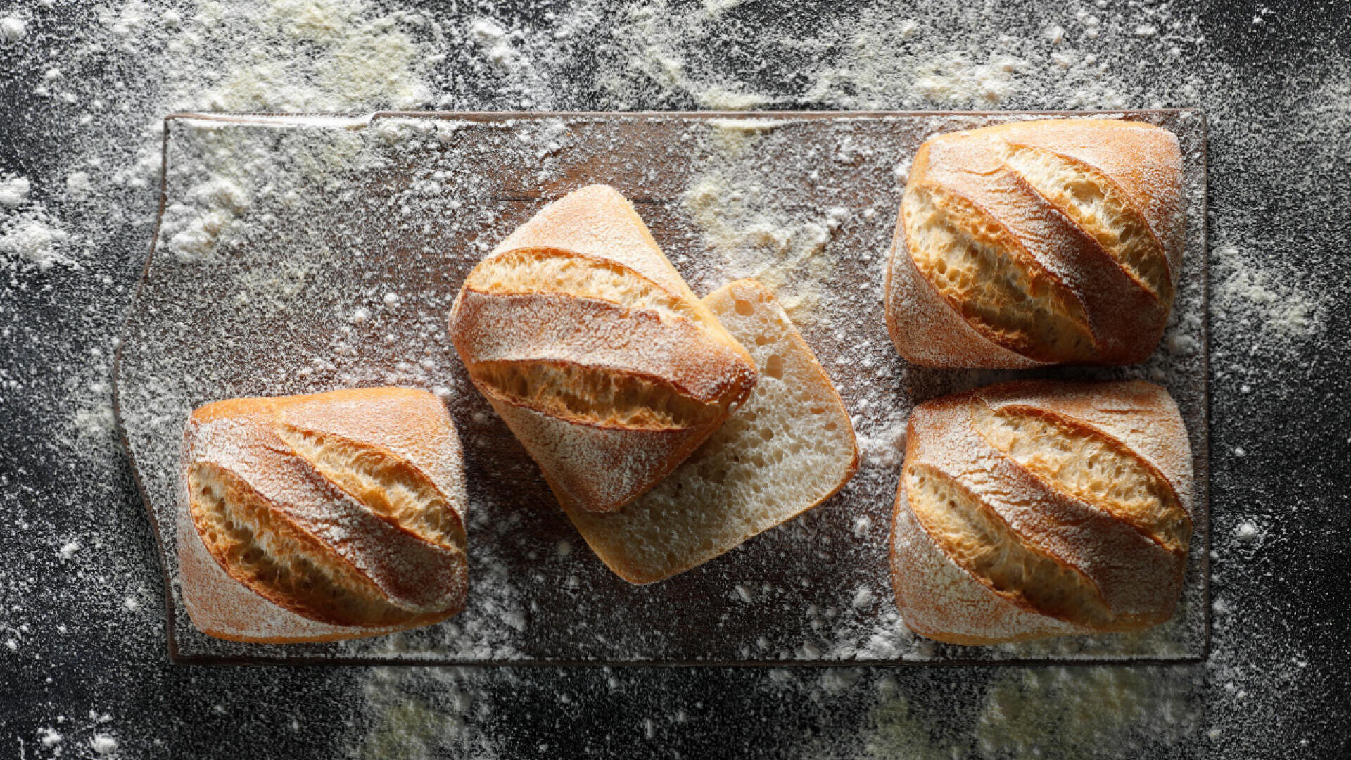 Vier quadratische, mit Mehl bestäubte Brötchen aus handwerklicher Herstellung werden auf einer bemehlten dunklen Oberfläche angeordnet, wobei ein Brötchen aufgeschnitten ist, um das weiche Innere zu zeigen.