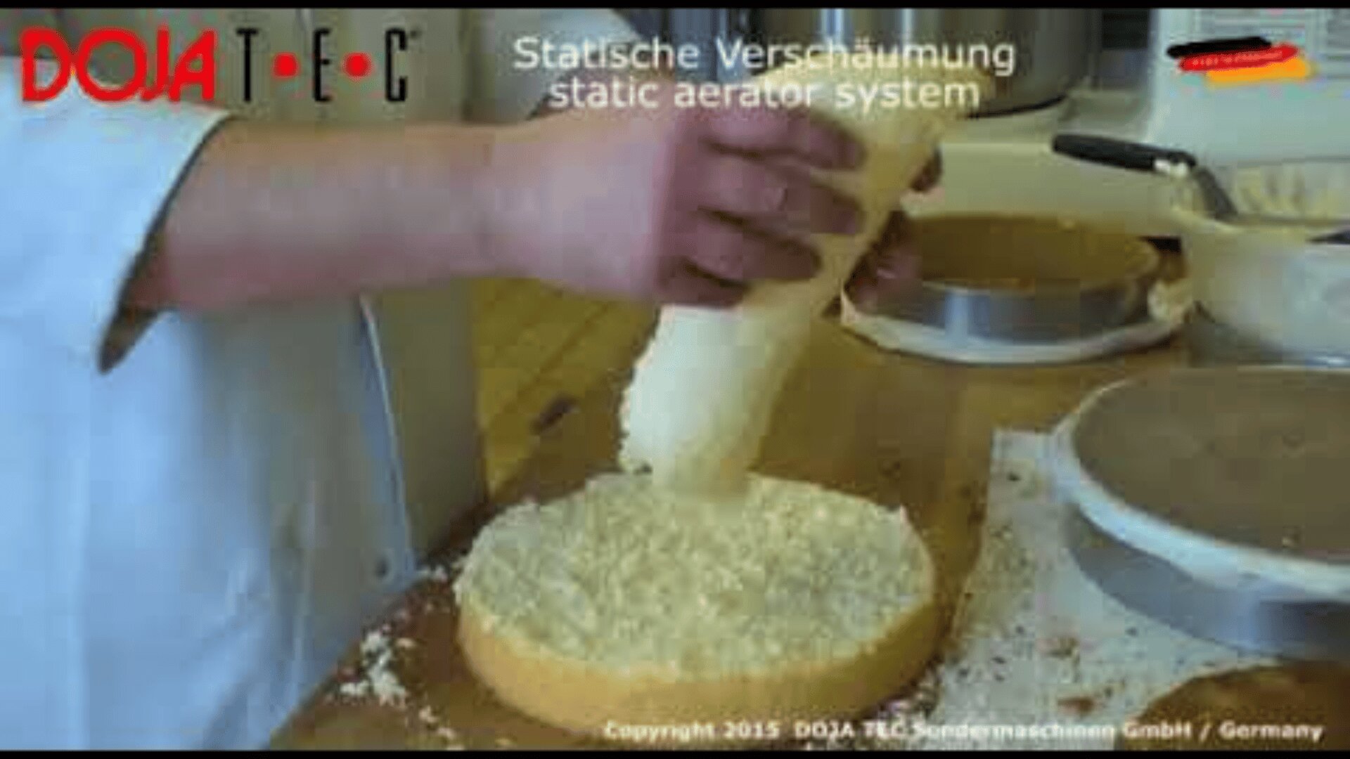 Eine Person verwendet ein zylindrisches Werkzeug, um in einer Küche eine schaumige Substanz auf einen runden Kuchenboden aufzutragen. Oben erscheint der Text Statische Verschäumung static aerator system.