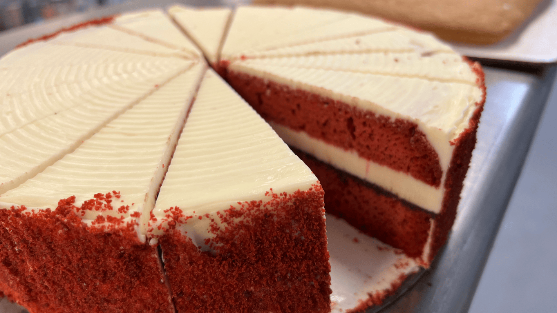 A close-up of a sliced red velvet cake with cream cheese frosting, showing two cake layers separated by frosting. One large slice is missing, revealing the inside texture. The cake sits on a metal surface.