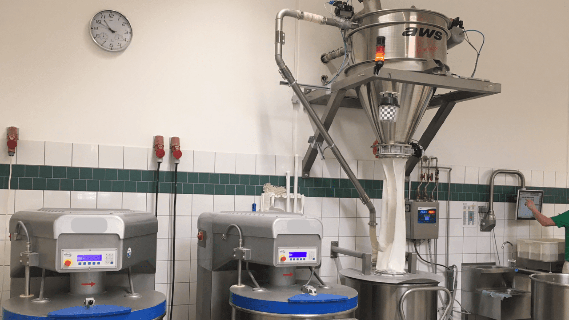 A commercial kitchen or food production facility with industrial machines, a large mixing or processing unit labeled “aws,” and a wall clock showing 9:16. A person’s hand is touching a wall-mounted device on the right.