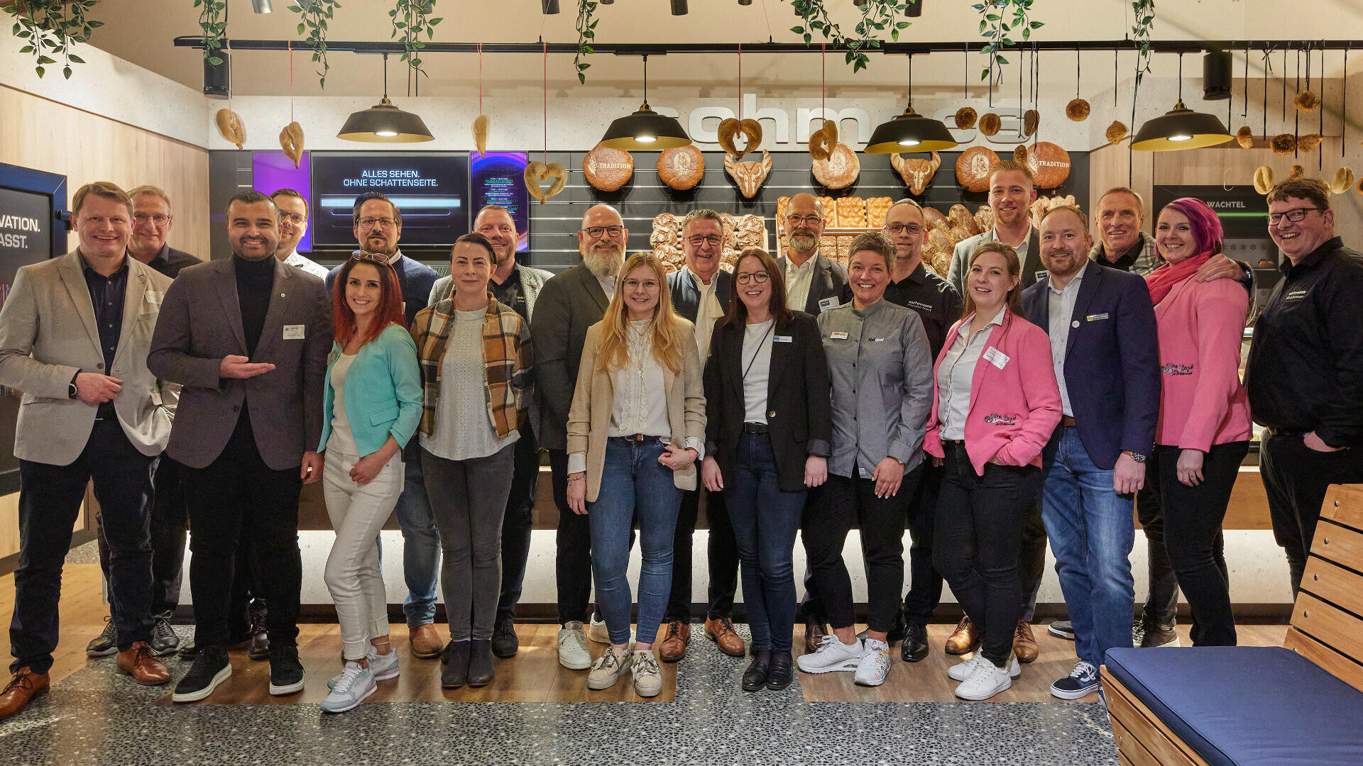 Eine Gruppe von 22 Personen, Männer und Frauen, steht in einem Innenraum zusammen und lächelt in die Kamera. Sie tragen Business-Casual-Kleidung, im Hintergrund ist ein modernes Café mit Holzdekor zu sehen.