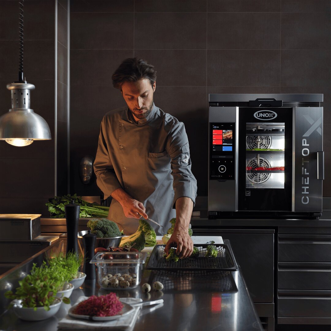 Ein Koch in grauer Uniform bereitet Gemüse auf einer Metalltheke in einer modernen, professionellen Küche zu. Im Hintergrund ist ein Industrieofen mit der Aufschrift CHEFTOP zu sehen, auf der Theke liegen frisches Gemüse und Kräuter.
