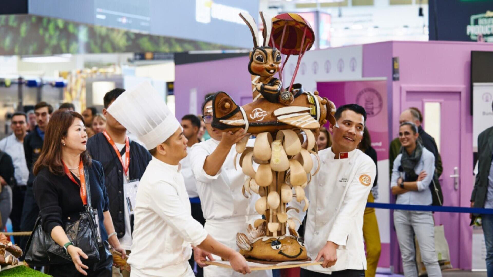 Three pastry chefs carry a large, elaborate chocolate sculpture of an insect character riding a flying machine while a crowd of onlookers watches at an indoor event or competition.