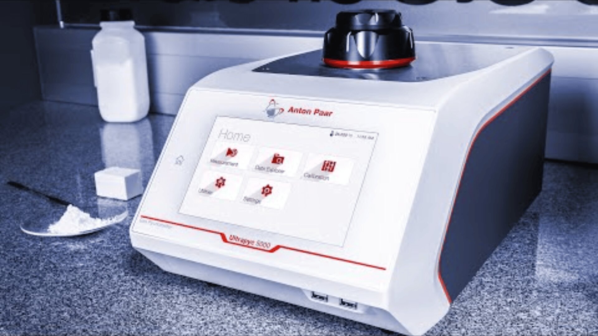 Auf einer Arbeitsfläche steht ein Laborgerät, das Anton Paar Density 5000. Ein Touchscreen-Display zeigt Menüoptionen an. Pulver, eine weiße Flasche und ein Würfel stehen daneben und deuten auf eine Probenanalyse hin.