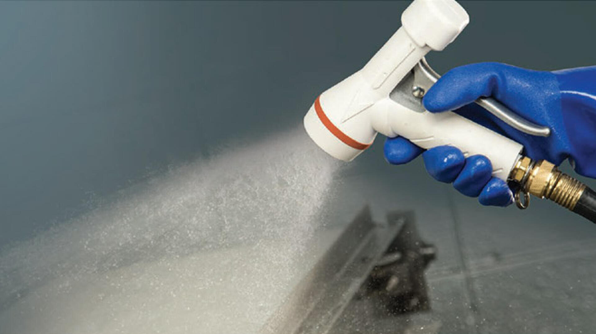 Eine Person mit einem blauen Handschuh sprüht Wasser aus einer weißen Schlauchdüse und erzeugt so einen Nebel auf einer Metalloberfläche.