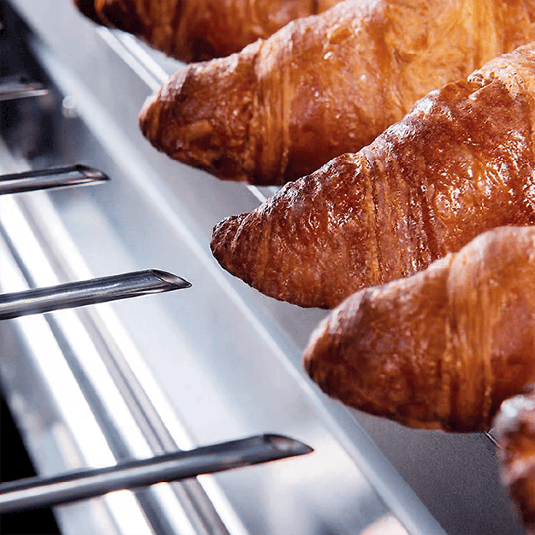 Mehrere goldbraune Croissants sind auf Metallstäben in einer Vitrine oder Bäckerei aufgereiht und zeigen ihre flockige Textur und glänzende, glasierte Oberfläche.