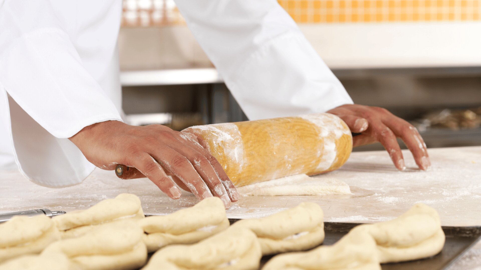 Eine Person im weißen Kochkittel benutzt ein hölzernes Nudelholz, um den Teig auf einer bemehlten Fläche zu glätten, wobei mehrere geformte Teigstücke im Vordergrund zu sehen sind.