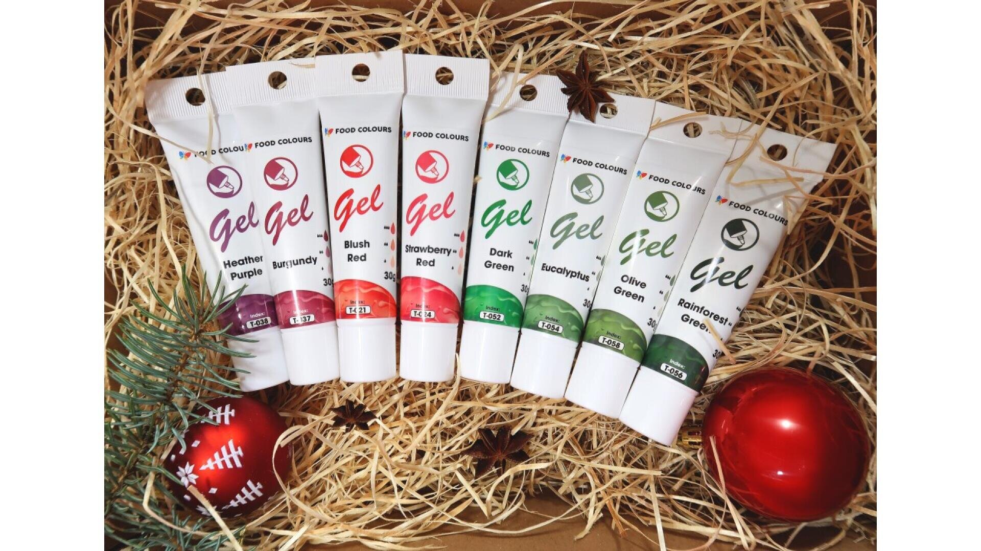 Sieben Tuben Gel-Lebensmittelfarbe in verschiedenen Rot-, Violett- und Grüntönen werden auf Stroh mit festlichen Dekorationen, darunter Tannenzweige und rote Ornamente, angeordnet.