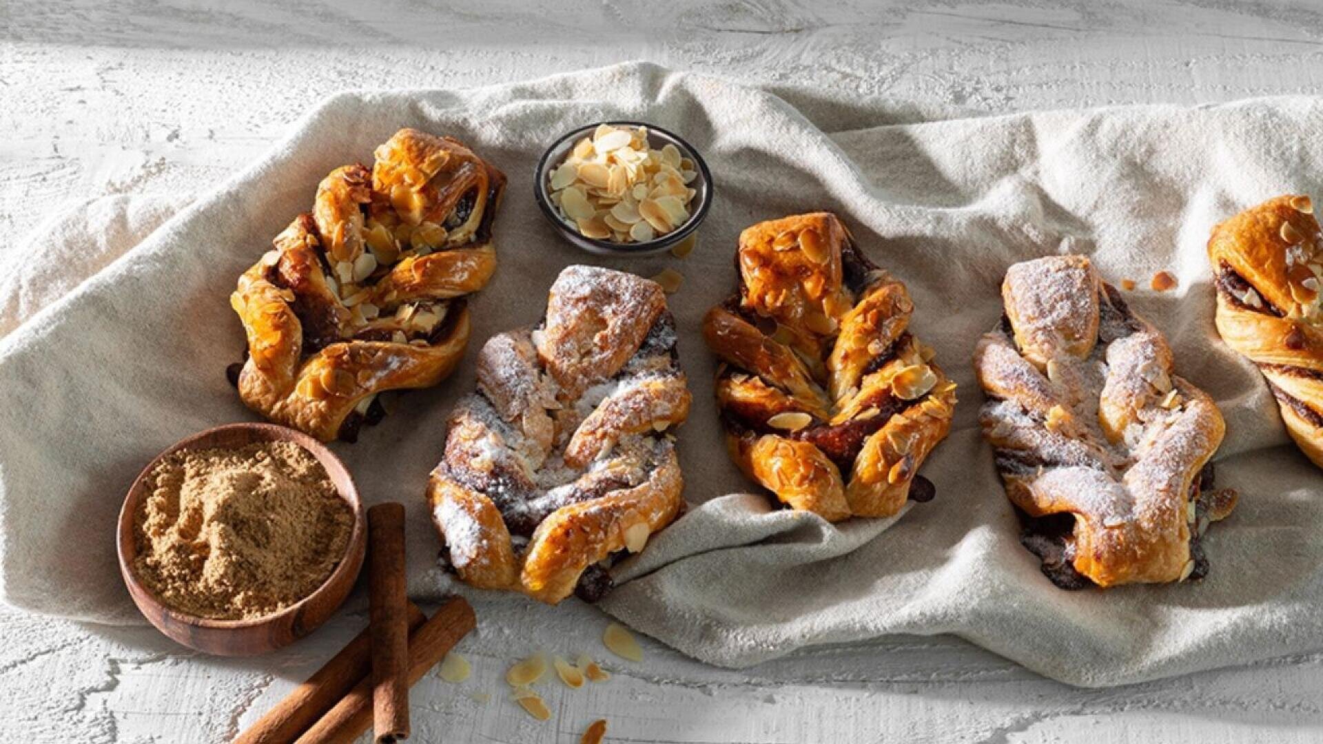 Four braided pastries dusted with powdered sugar are arranged on a beige cloth. A small bowl of sliced almonds, a bowl of brown sugar, and two cinnamon sticks are beside them on a rustic white surface.
