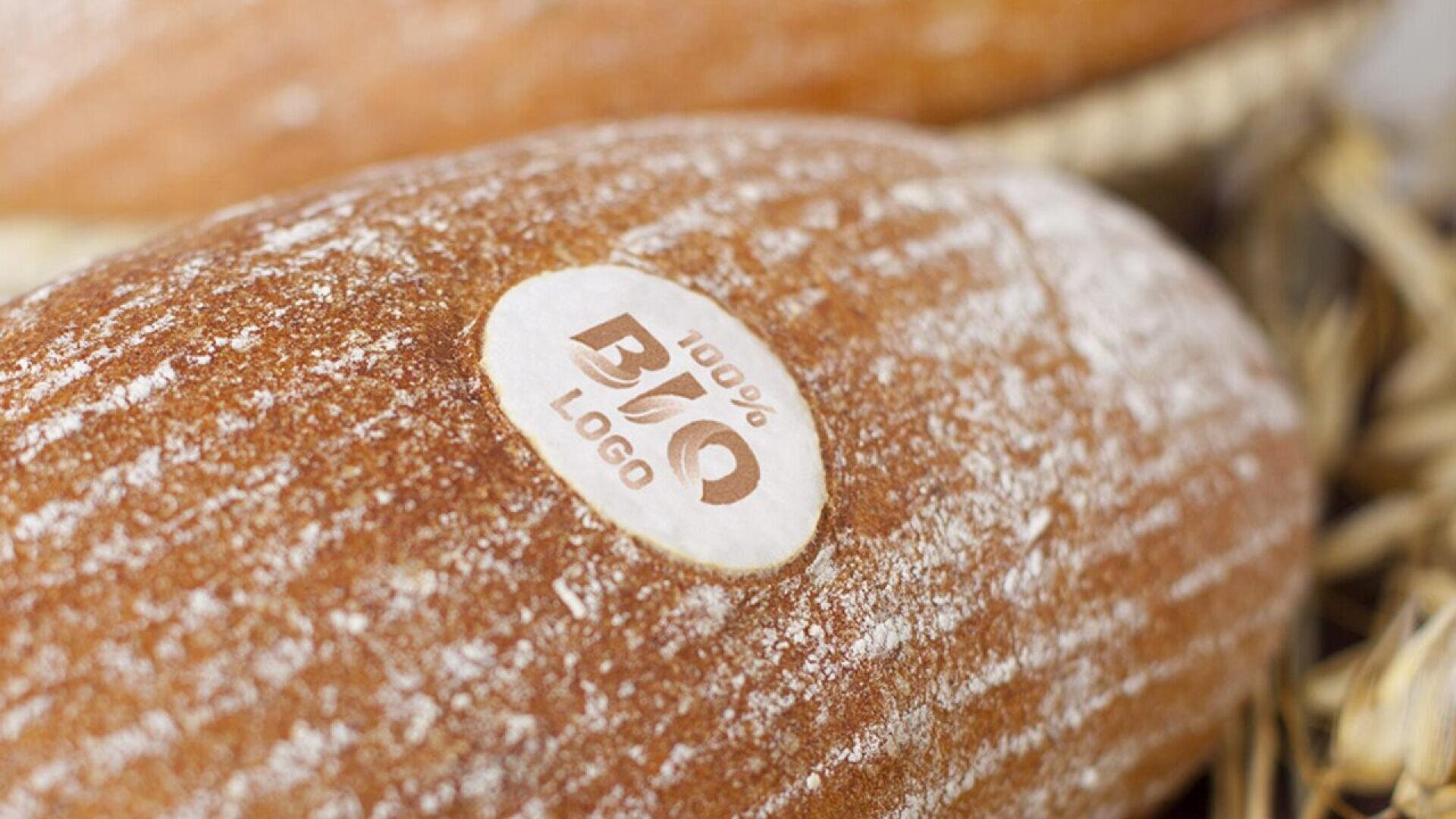 Nahaufnahme eines mit Mehl bestäubten Brotlaibs, auf dem ein runder weißer Aufkleber mit der Aufschrift "100% BIO LOGO" zu sehen ist. Ein weiterer Laib und etwas Stroh sind im unscharfen Hintergrund zu sehen.
