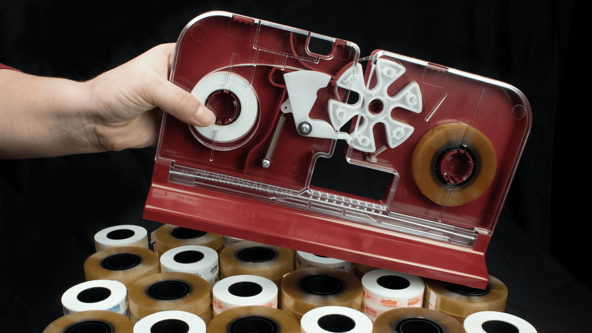 A hand holds a red tape dispenser with clear and brown rolls of tape loaded inside, above a table covered with many rolls of packaging tape against a black background.