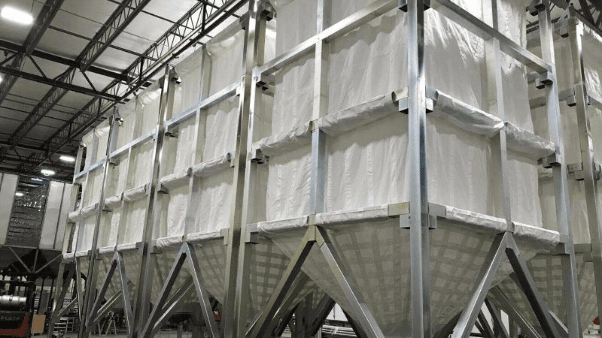 Große industrielle Stoffsilos mit Metallrahmen stehen in einer Reihe in einer Lagerhalle. Die Silos haben konische Böden für die Ausgabe von Materialien, und der Bereich ist mit einer hohen Decke und sichtbaren Metallträgern gut beleuchtet.