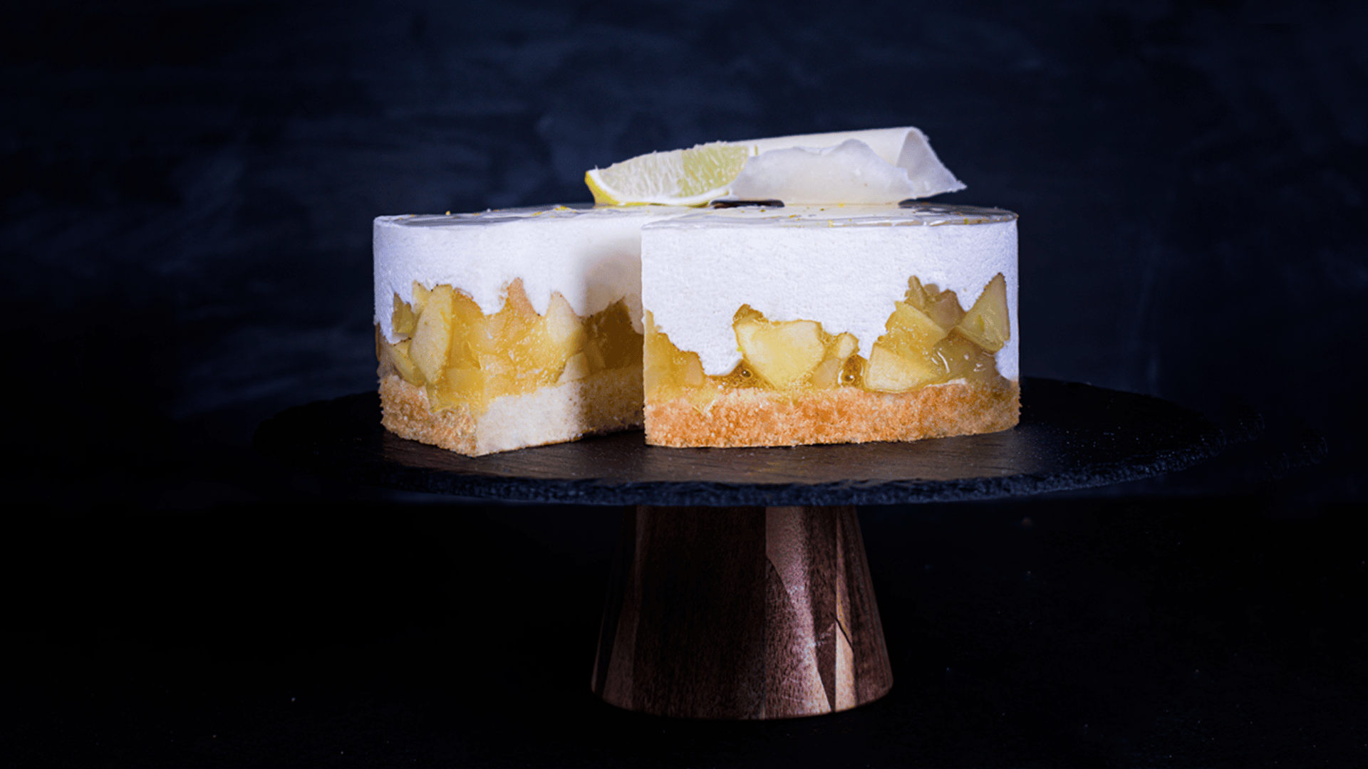 A round layered dessert with a golden crust, a middle layer of diced fruit, and a thick, creamy white topping, sits on a dark cake stand. A lemon wedge garnishes the dessert. The background is dark.
