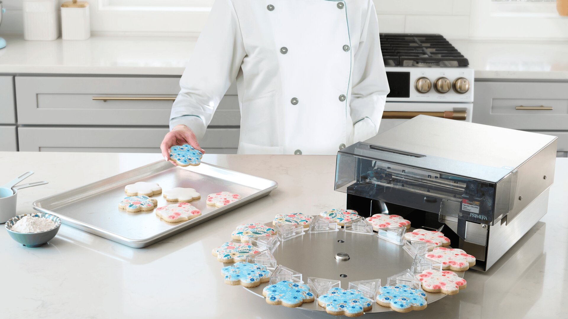 Ein Bäcker im weißen Kochkittel hält einen dekorierten Keks in einer modernen Küche. Auf dem Tresen stehen ein Keksdrucker und ein rotierendes Tablett mit Keksen, von denen einige mit blauer und rosa Glasur verziert sind.