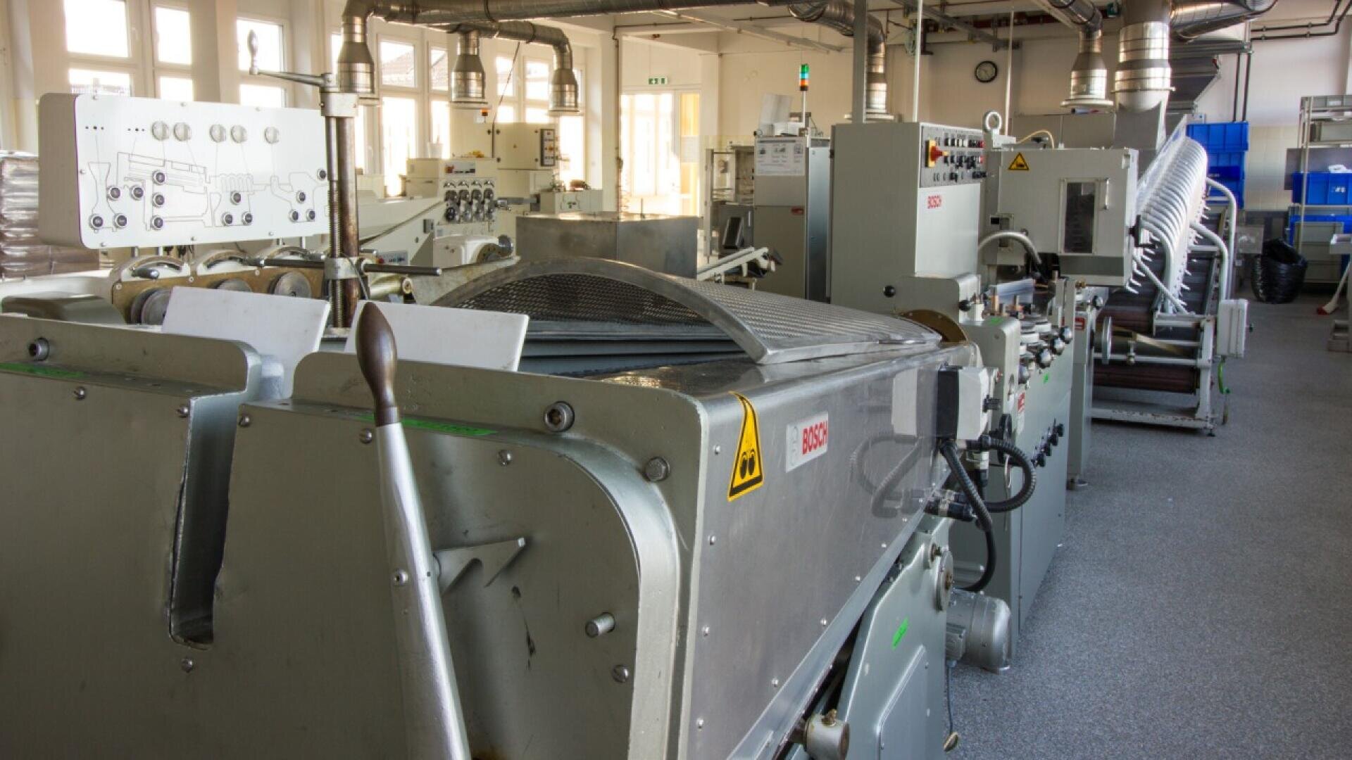 Industrial machines with control panels and levers are arranged in a bright, spacious factory room with large windows and ventilation pipes on the ceiling. The scene suggests a manufacturing or processing facility.