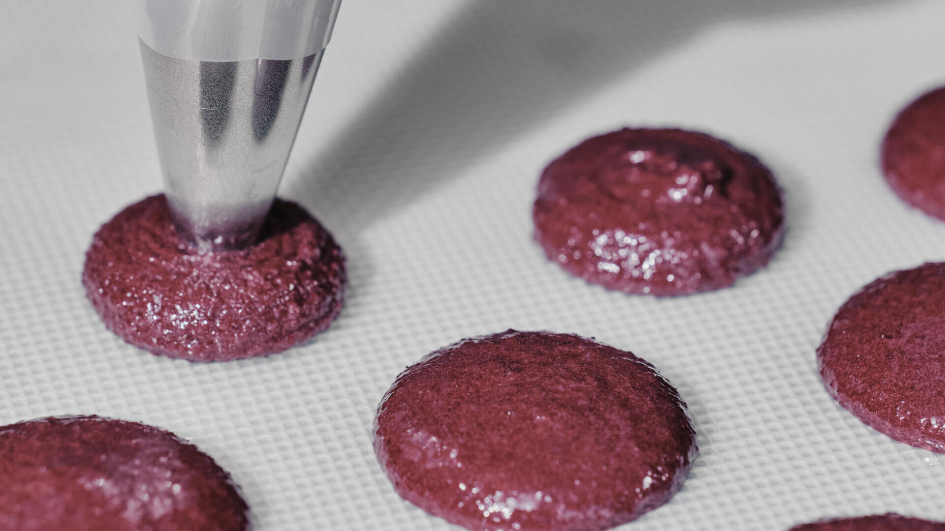 A piping bag squeezes purple macaron batter onto a baking mat, with several round, glossy macaron shells already piped nearby.