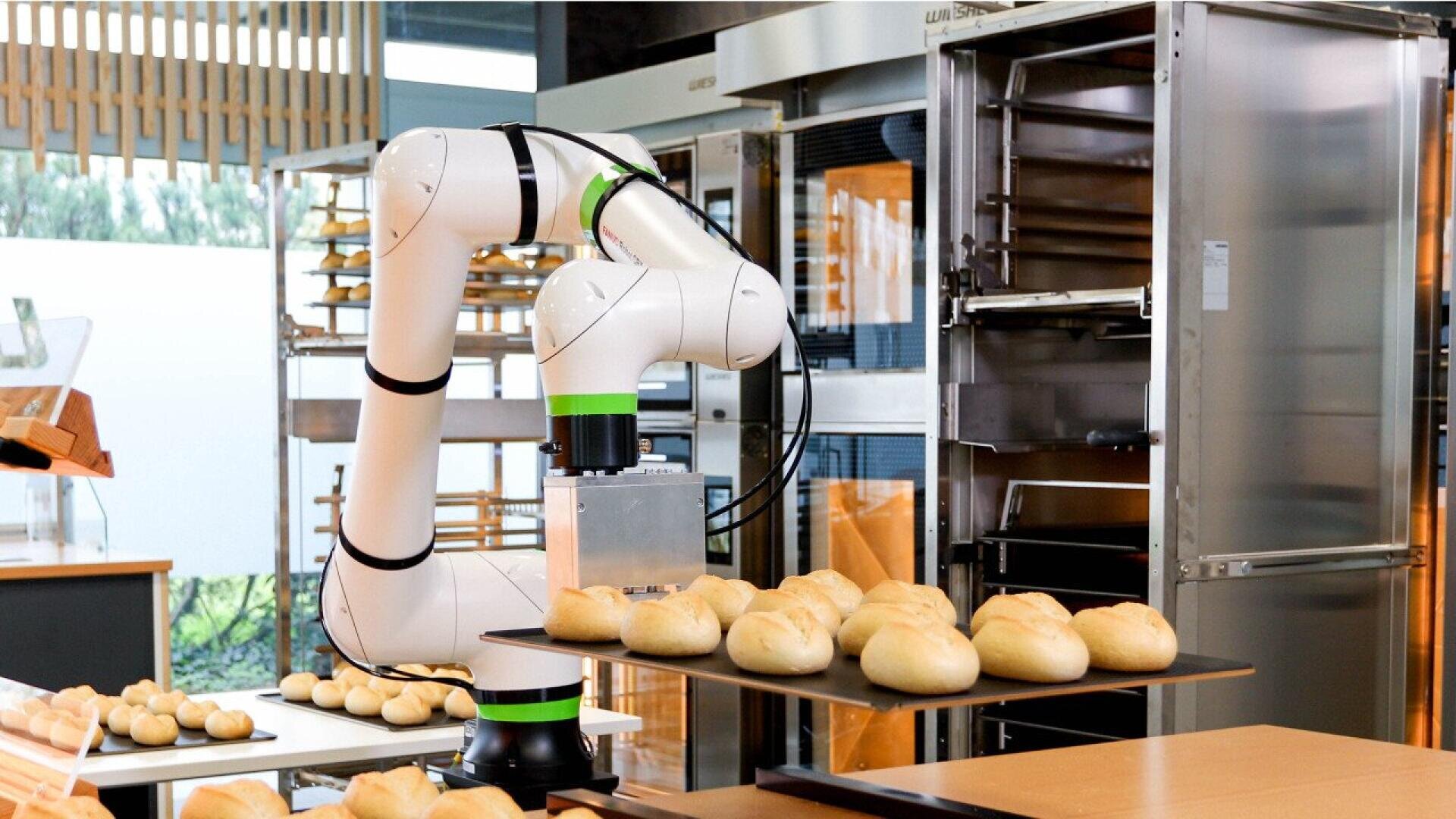 A robotic arm places freshly baked bread rolls onto a tray in a modern bakery, surrounded by shelves and industrial baking equipment.