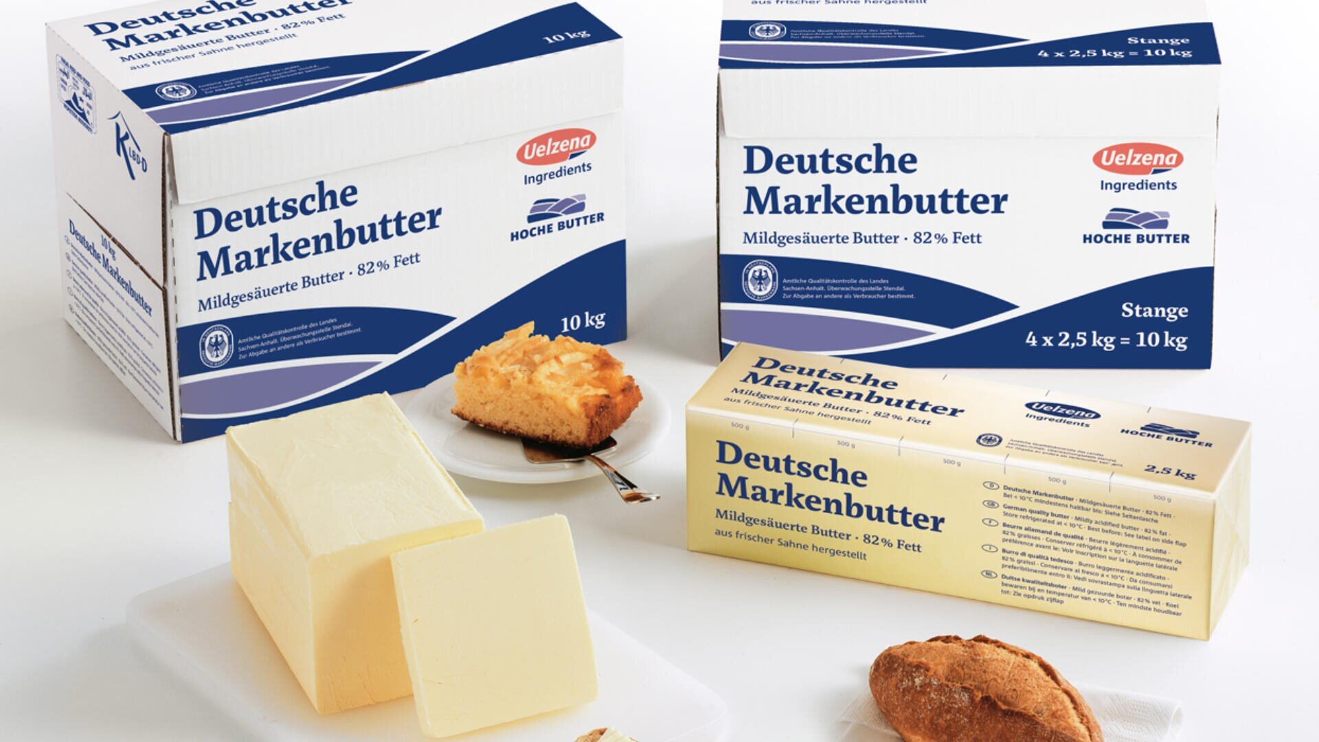 Schachteln und ein Block Deutsche Markenbutter werden mit einer Butterdose, einer Scheibe Brot und einem Stück Kuchen auf einer weißen Fläche präsentiert. Auf der Verpackung ist angegeben, dass die Butter 82 % Fett enthält.