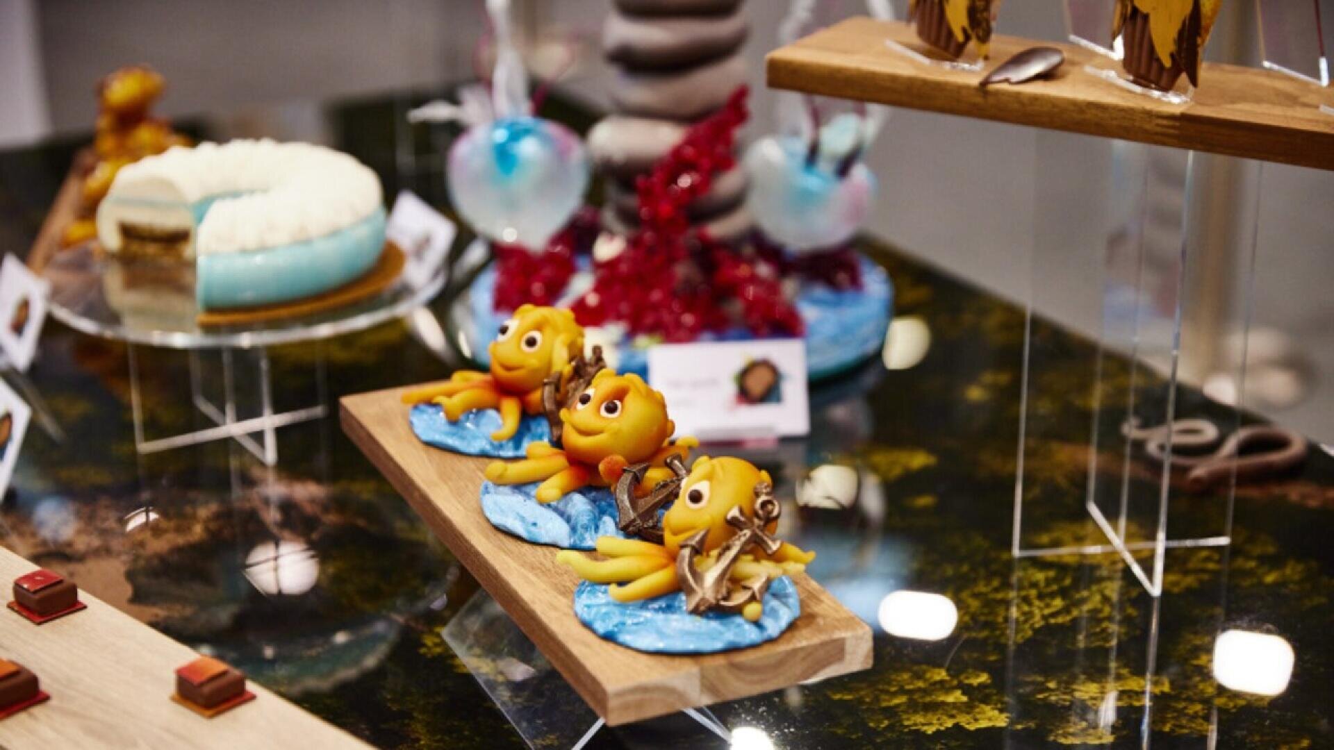 Three fish-shaped pastries with expressive faces sit on a bed of blue decoration, displayed on a wooden tray. Other artistic desserts and confections are visible in the background on a glass table.