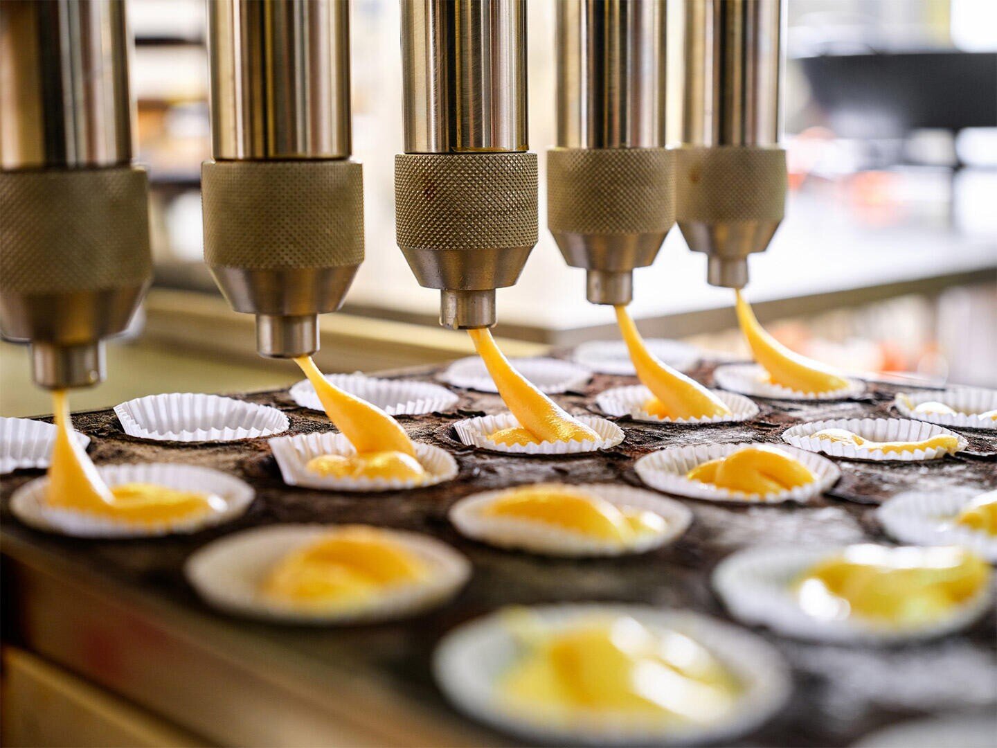 Industrial baking machine dispenses yellow batter into rows of white paper cupcake liners, preparing multiple cupcakes at once in a factory setting.