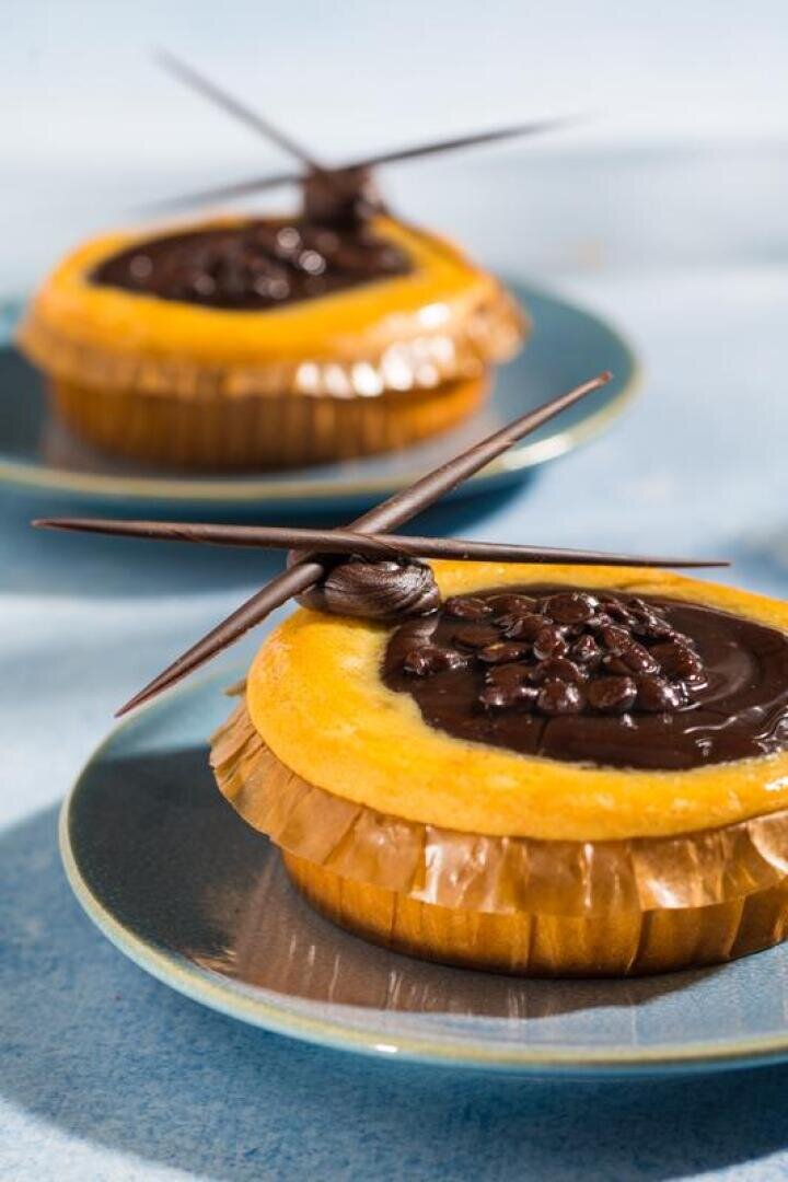 Two small cheesecakes topped with glossy chocolate sauce and decorative chocolate sticks, each served on a blue plate with a ruffled paper liner.