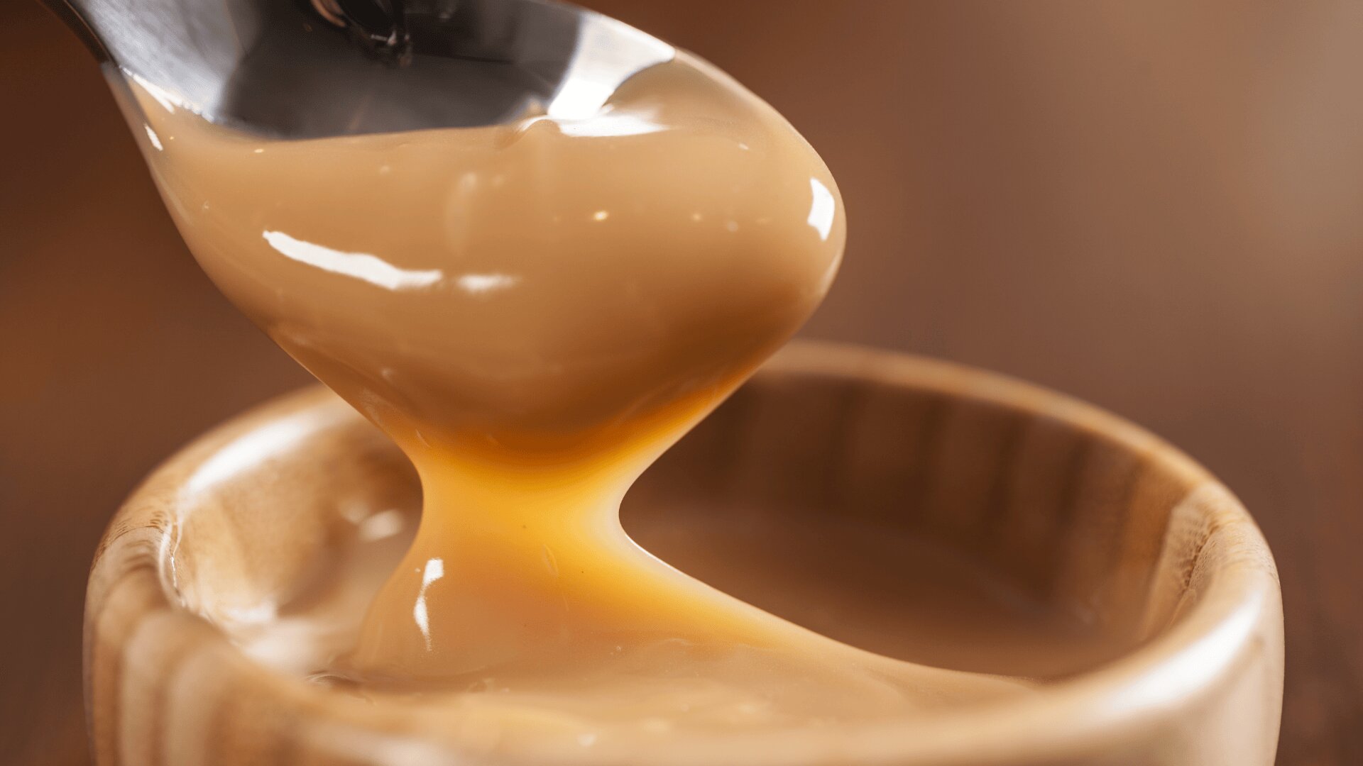 A close-up image of a spoon lifting thick, creamy caramel sauce from a small wooden bowl, with the sauce smoothly dripping back into the bowl.