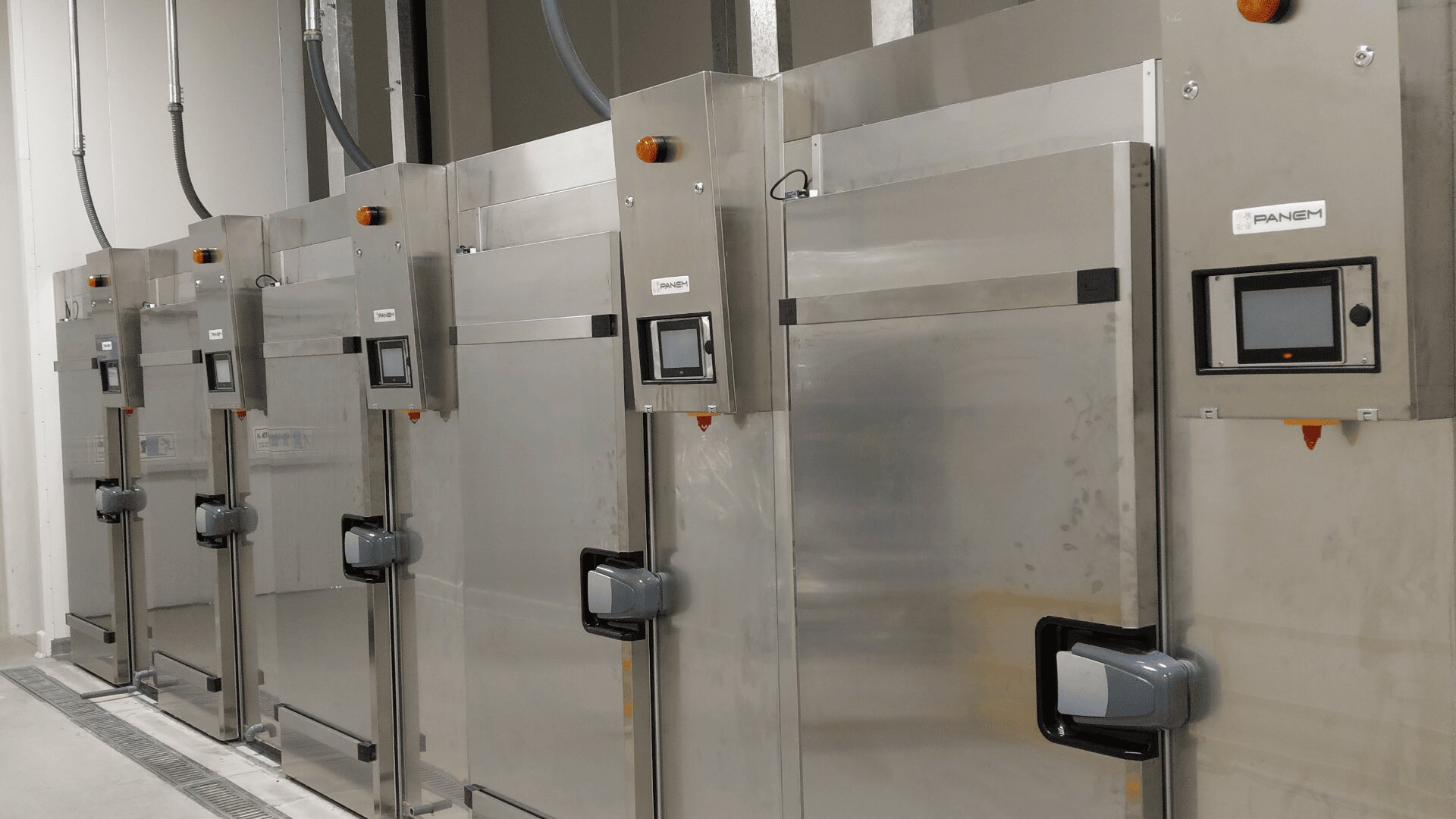A row of large, stainless steel industrial ovens or proofers with control panels and handles, lined up against a white wall in a clean, modern facility.