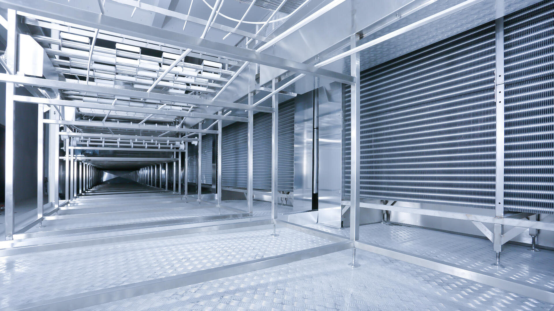 Interior view of a large, industrial metal air duct or ventilation system with silver panels, metal framework, and gridded flooring, showing clean, modern construction and a sense of depth.