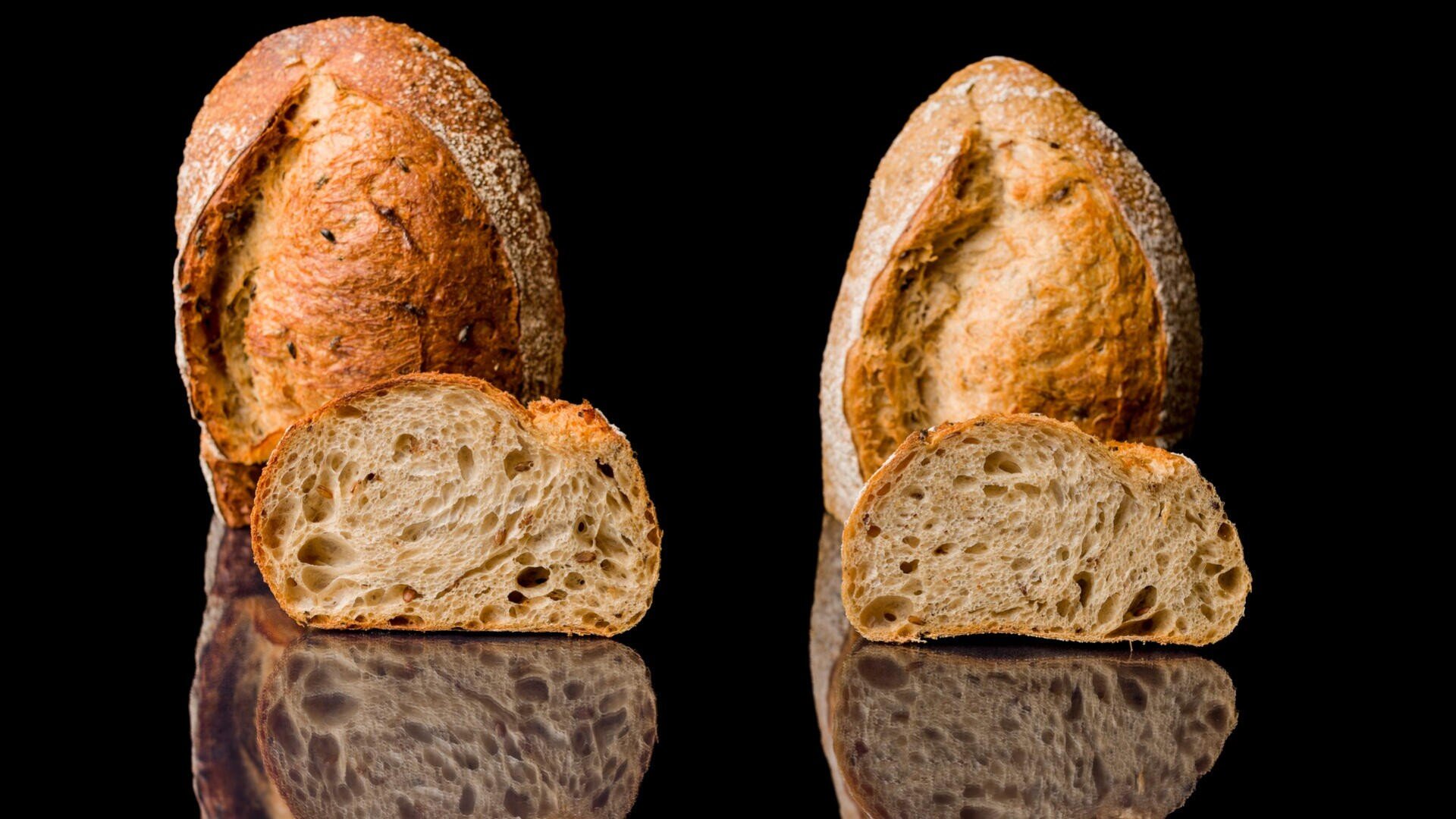 Two round loaves of rustic bread with a slice cut from each, displaying airy, textured interiors, are set against a black background with their reflections visible below.