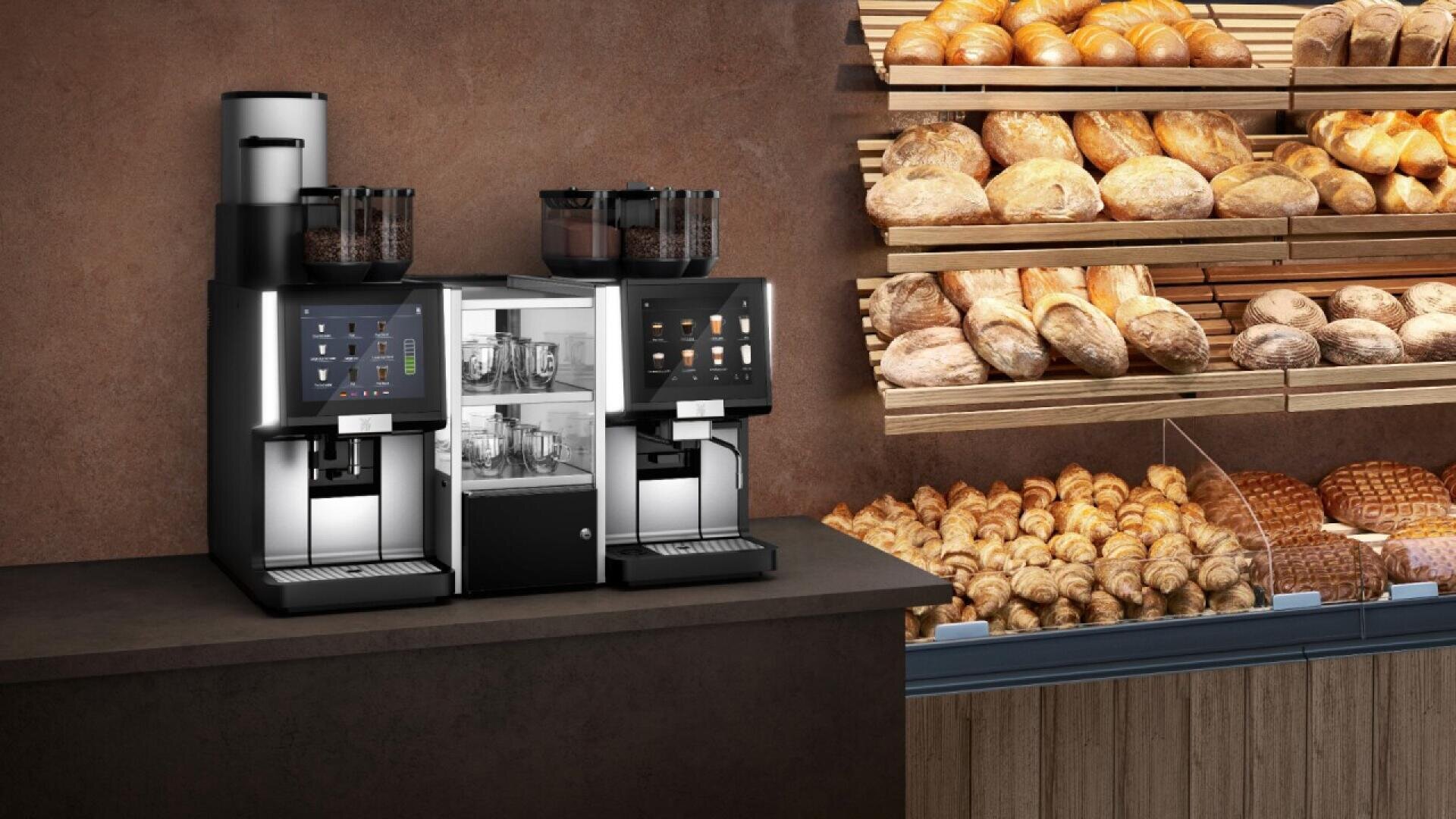 Two modern coffee machines on a counter beside shelves filled with a variety of breads and pastries in a bakery setting.