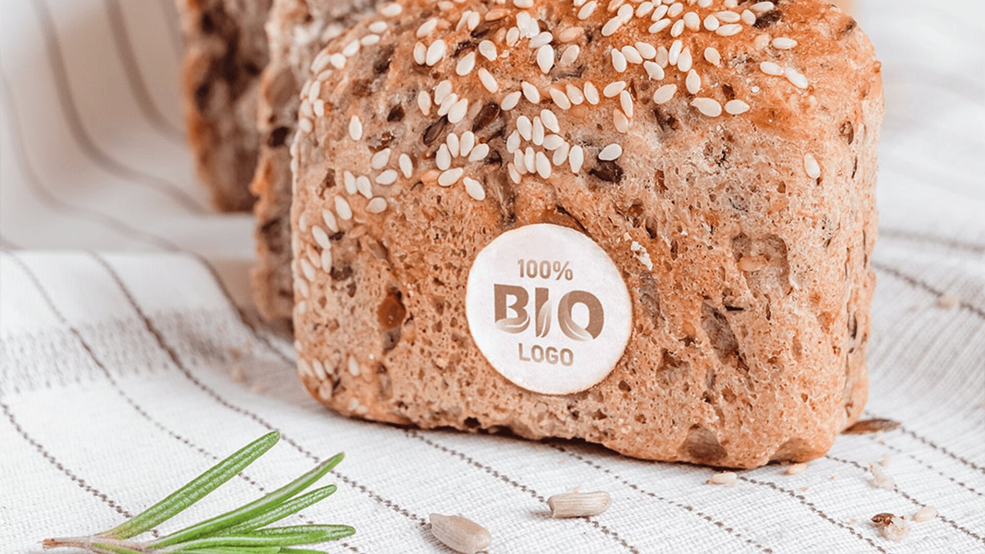A loaf of multigrain bread topped with sesame seeds sits on a striped cloth. A round label on the bread reads “100% BIO LOGO.” A sprig of rosemary and a few sunflower seeds are in the foreground.