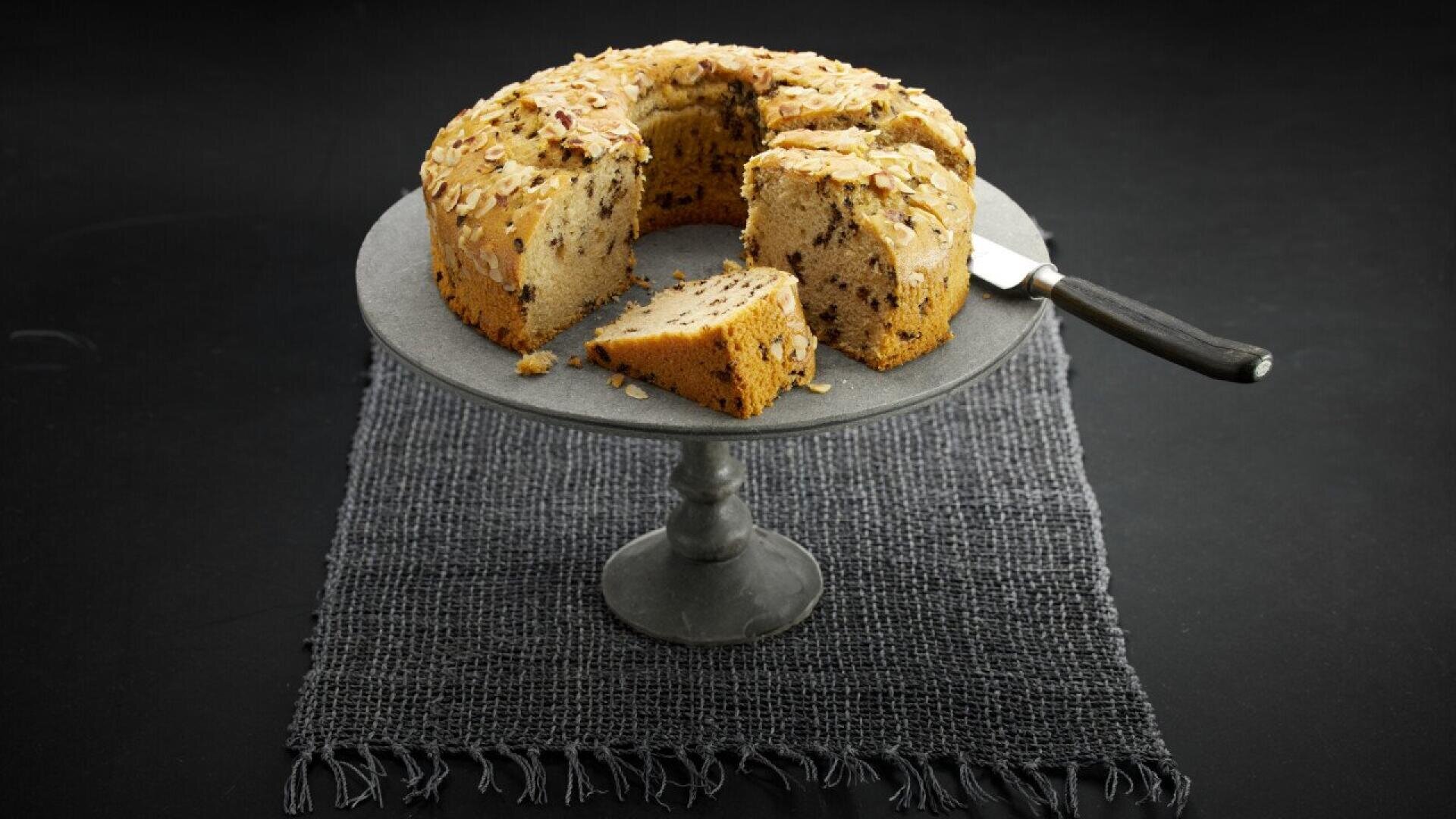 A round, nut-topped cake with a slice cut out sits on a gray cake stand over a dark textured mat. A knife rests beside the cut slice on the stand. The background is black.