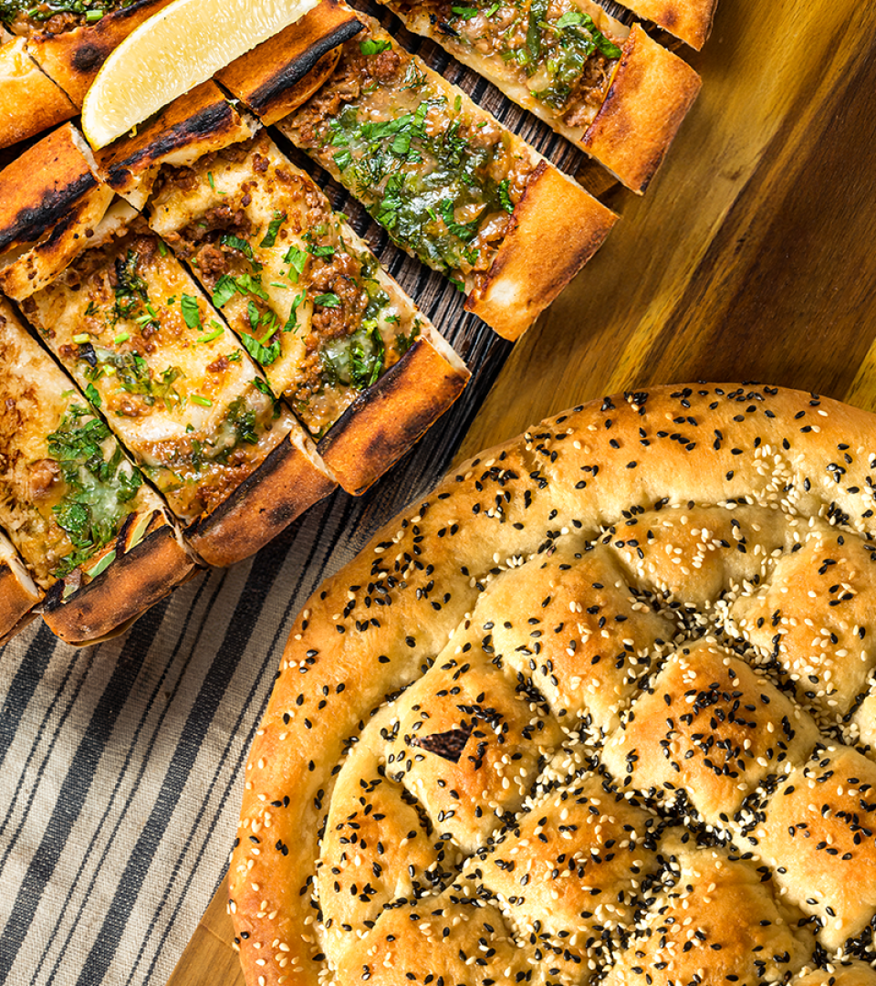 Close-up of Turkish pide slices topped with herbs and minced meat, next to a round loaf of sesame and black seed covered bread on a striped cloth and wooden surface.