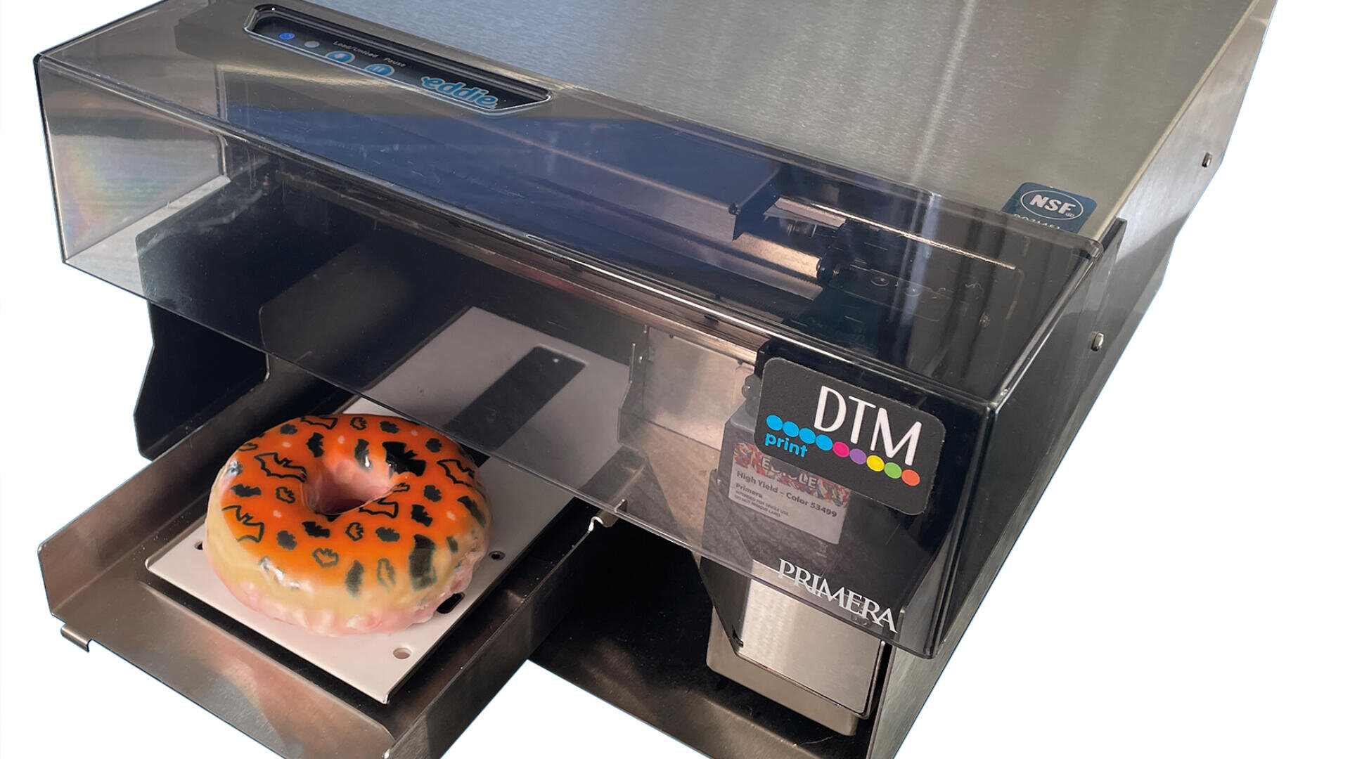 Ein Donut mit orangefarbenem und braunem Zuckerguss wird in einem kommerziellen Lebensmitteldrucker mit den Bezeichnungen DTM und PRIMERA bedruckt oder dekoriert. Der Drucker hat eine transparente Abdeckung und ein Metallgehäuse.