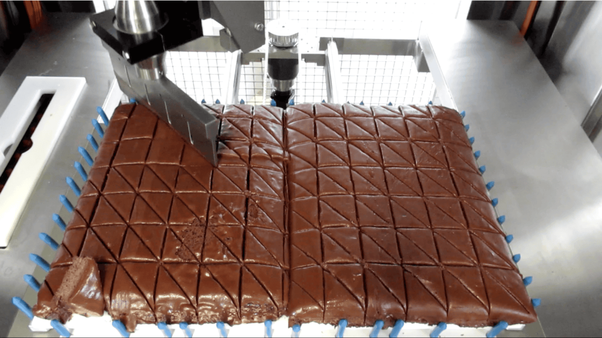 Eine Maschine mit einer Metallklinge schneidet einen großen rechteckigen Schokoladenkuchen auf einer Metallfläche in rautenförmige Stücke. Blaue Plastikstifte säumen die Seiten des Kuchens.